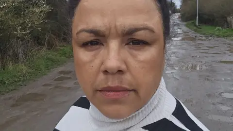 Kerry Watson looking at the camera. She's standing outside on an unadopted road with lots of puddles, and wearing a black and white polo neck jumper