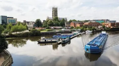 The photo shows a wide stretch of river with a blue barge in the forefront and a number of smaller barges in the background. Doncaster Minister overlooks the water