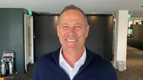 Photograph of Simon Ismail from the developer Salboy. He has short grey hair and is wearing a white shirt and navy blue jumper with a zip which is opened below the neck. The image shows him inside the company's head office on Salford Crescent. He is smiling.