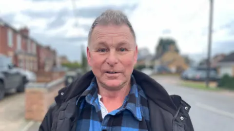 Laura Foster/BBC Danny Jones with short grey hair looking at the camera and wearing a blue shirt and a blue anorak, standing next to a road with two-storey houses on the left and a low brick wall. The sky is overcast.