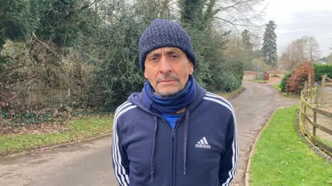 A man in a hat and scarf stands outside on a country road with a serious expression on his face. He is wearing a blue Adidas top