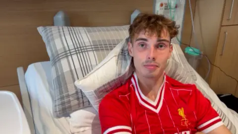 Mousumi Bakshi/BBC A young man wearing a red LFC football top is sat in a hospital bed and looking directly at the camera