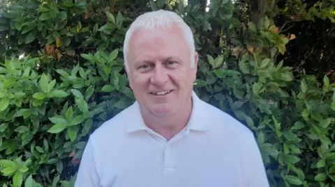 Derek McGilligan has short grey hair and is wearing a white polo shirt. He is standing in front of a large green bush outside.