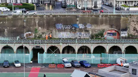 A wide drone shot of Madeira Terrace, showing the area fenced off from the public, two workers in hi vis standing on top of the terrace and several cars parked in front of the structure.