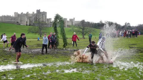 PA Media Two players clash with a large spray of mud around them. Other players are waiting to see where the ball comes loose.