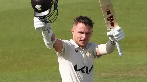 Sam Curran raises his bat and helmet after reaching 100 for Surrey against Durham