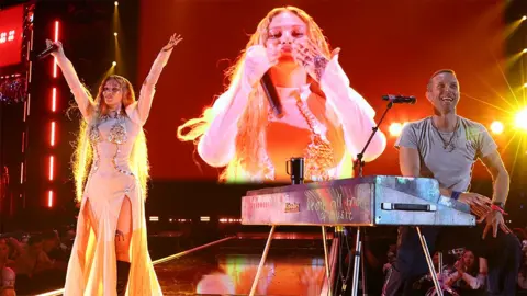 Getty Images Ileana and Coldplay's Chris Martin perform on stage at the 2024 iHeartRadio Music Festival in Las Vegas. Ileana wears a long, light-colored outfit and extends her arms toward the sky. Her image is on the big screen behind her, with Chris Martin sitting in front of a silver keyboard, wearing a gray T-shirt, smiling and looking out at the audience.