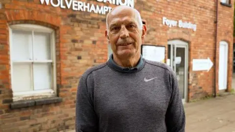 A man with short grey hair on the sides of his head is looking at the camera and smiling. He is wearing a grey Nike jumper and is standing in front of a red brick building.