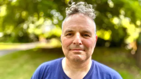 Martin Giles/BBC Mark Evans stares at the camera with a neutral expression. He wears a dark blue t-shirt. There is a blurred background with trees, grass and a path