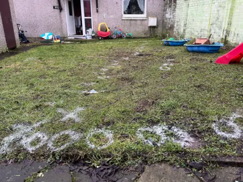 A general view of the back garden of the house at Ancrum Court. The grass has white writing spray painted on it containing letters and numbers.