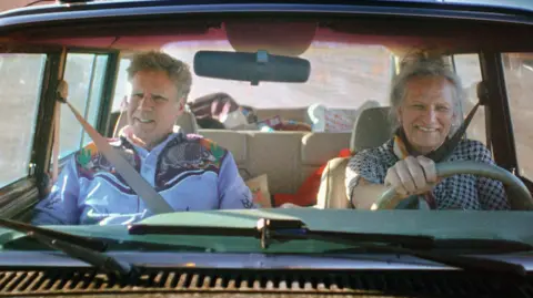 Netflix Will Ferrell and Harper Steele in a car driven by Harper. He is wearing a blue cowboy shirt and she is wearing a black gingham top.