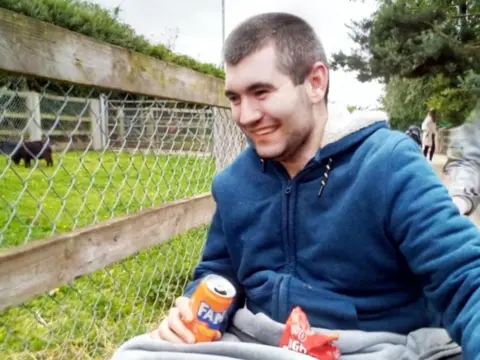 Aaron on a trip to a farm. He is smiling and holding a can of fizzy drink. He is sitting in a wheelchair with a blanket over his knees. In the background there is black goat grazing on grass behind a fence.
