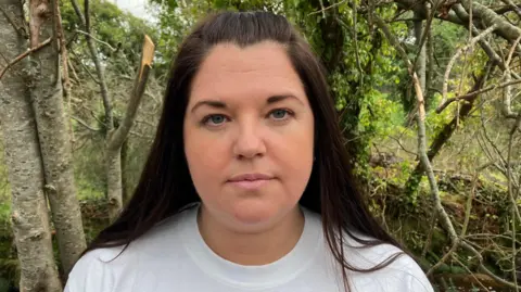 Gail has a white T-shirt on and long brown hair, some of which is tied back. She is standing in front of some trees.