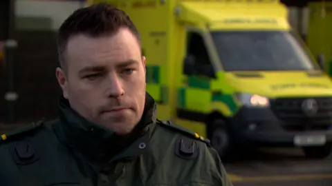 Paramedic Johnny Hanna. He has short dark brown hair and blue eyes and wearing an ambulance uniform. And ambulance can be seen in the background.