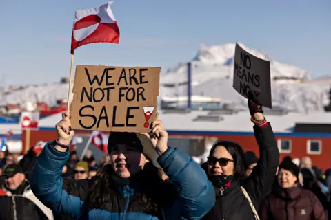 AFP via Getty Images A protester holds a placard reading 'We are not for sale', with other protesters behind them against a backdrop of snowy mountains and the Greenland flag.
