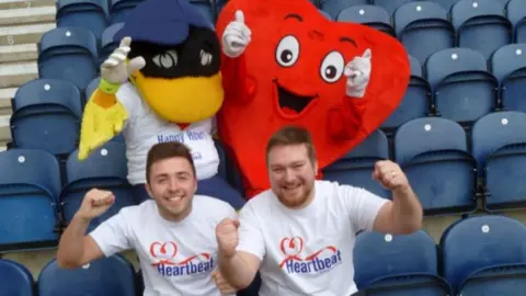 GOFUNDME An image showing two men, in white tshirts, sat on blue seats. Behind them is a red heart mascot and a duck mascot.