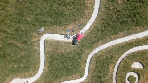 BBC/ Kevin Church A man with a red top and a hat is seen from above. He is on his fours over a white chalk line with his spade and a plastic bag next to him. Other parts of the white outline of the giant are seen including a circle - presumably one of his eyes, a semicircle - probably his eyebrow and a wriggling line.