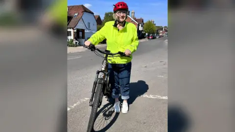 Rosemary Shrager in jeans, a bright yellow high vis jacket and a bike helmet. She is standing on a road next to a bike.
