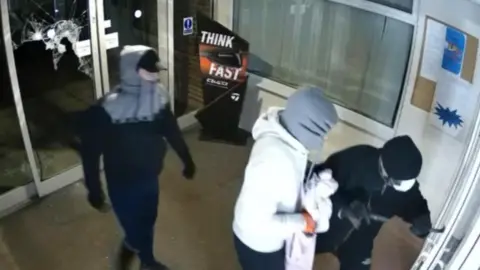 CCTV shows three people wearing hoods and covering their faces in the entrance to the golf club at West Malling. The club has glass doors and internal windows with blinds. It is night-time outside. The people shown on the CCTV are using a crowbar to get through internal glass doors. A door behind them has broken glass.