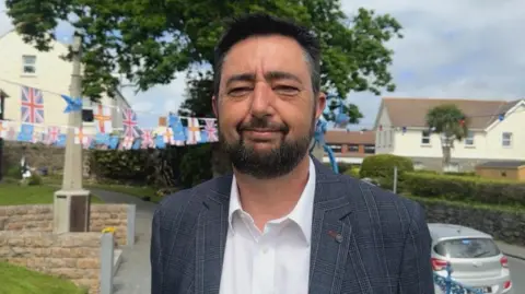 BBC Ricky Mahy looks at the camera, he is wearing a white shirt and checked jacket