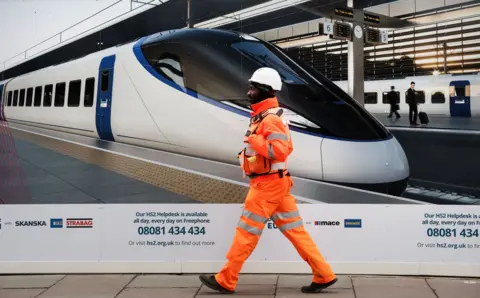  EPA-EFE/REX/Shutterstock A High Speed 2 worker dressed in orange hi-vis passes a billboard of a HS2 train at the Euston terminal.