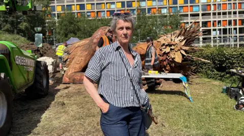 Simon Thake A woman with short grey hair stands in the sunshine, hands in her pockets. Behind her a large wooden sculpture rests on the grass.