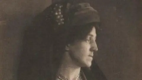 The William Morris Society A lack and white image of a woman - May Morris - with her head turned to to left-hand side. She is wearing a head piece with long material at the back. 