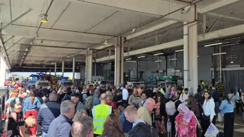 David Evans Dozens of passengers stand outside under a roof covering with airport staff in fluorescent bibs. Fire engines can be seen at the rear of the crowds.