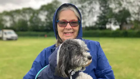 JOANNA TAYLOR/BBC Jan Forster smiling at the camera and holding a small black-and-white dog. She is wearing a blue raincoat with the hood up and tinted glasses with brightly coloured frames. behind her is the field, fence and a white van. 