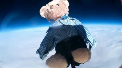 Walhampton School Small, brown bear, wearing school uniform, navy blue shorts and a blue and white checked shirt flying attached to a weather balloon above clouds
