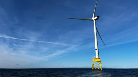 A single turbine in the foreground with several others further away in an offshore wind farm against a blue sky.