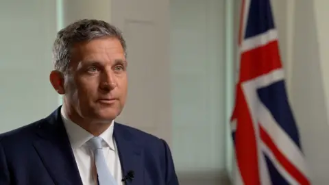 BBC Prisons minister James Timpson is pictured on the left hand side of the frame wearing a navy blue suit jacket, white shirt and pale blue tie. A Union Jack flag can be seen on the right hand side of the frame.