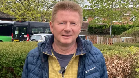 Rob Mariner from Blandford. His wife Mary has Alzheimers disease and uses the centre twice a week. He is standing next to a hedge with cars behind and a row of homes and trees. He is wearing a yellow jumper and a blue coat. 