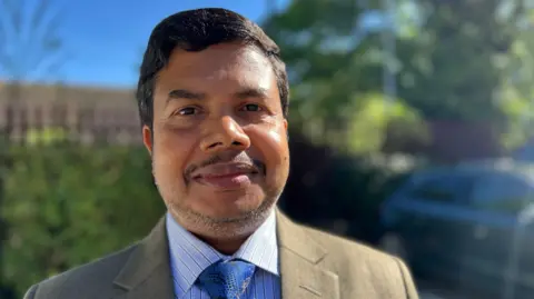 Head and shoulder image of Delowar Hossain. He has dark hair, stubble and is wearing a beige jacket, blue checked shirt and dark blue tie. He is smiling at the camera and standing in front of a car. 
