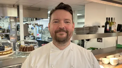 Carla Fowler/BBC Jon Smith smiling at the camera with an industrial kitchen behind him. He is in chef whites and has a brown beard and brown hair.