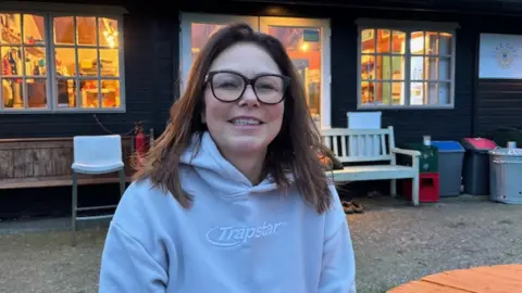 Rachel in a midshot, smiling to camera while sitting on a picnic table in front of a hut-style building at Court Farm Lakes. She is wearing a grey hoodie and has long brown hair and black-rimmed glasses. 