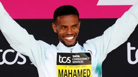 Mahamed Mahamed holding his arms aloft, smiling. He is wearing a white, long-sleeved top. He is standing in front of pink and black banners for the tcs London Marathon. He is wearing a yellow and black sign on his chest saying 'Mahamed'.