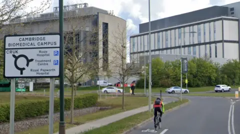 Google Man cycling on a bike past a roundabout sign with Cambridge Biomedical Campus written on the top of it