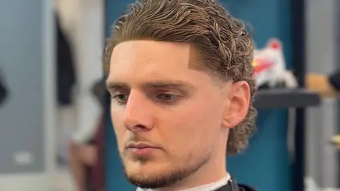 Musaab Somobwane A man wearing a barber cape sits in a chair, showing a close‑up of a haircut, with a sharp line delineating his otherwise wavy light brown hair from his forehead.
