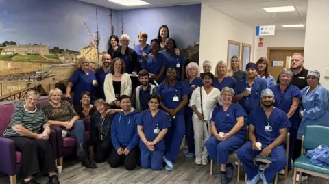 Royal Cornwall Hospitals NHS Trust A group of the team stood and sat together. They are all looking the camera and smiling. 