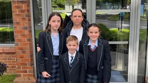 Three children in school uniforms look at the camera. Standing behind them is their mother who has her hand on their shoulder. They are standing outside their front door.