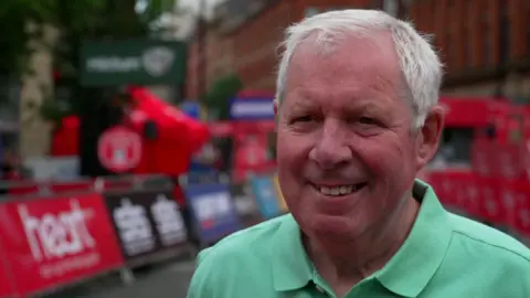 A man with short grey hair and a mint coloured polo shirt is smiling as he is interviewed beside the route of the race.