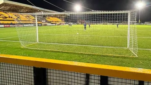 A view from behind the goal looking onto a football pitch, where a few players are warming up. On the left is an empty stand with yellow and black seats spelling out SFC. 