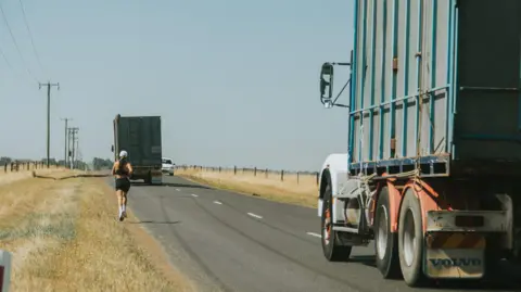Megan Chapple Megan Chapple running in Tasmania. Two trucks are seen passing her on a clear day.