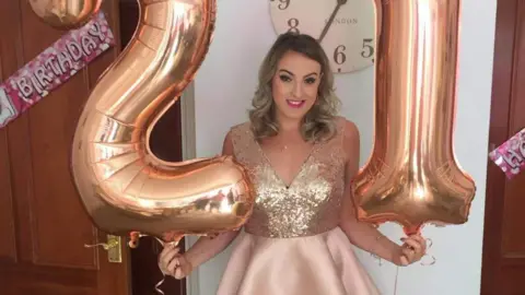 Family photo Bethan holding two balloons in the shape of a 21. She is wearing a dress with a sparkly top and a light pink skirt, and is wearing pink lipstick. Her hair is curled. There are pink birthday banners on two doors either side of her.