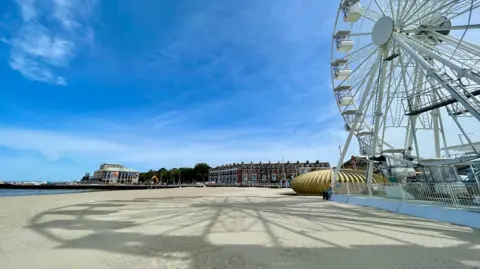 Rio South West The sandy beach with the shadow of a ferris wheel on its surface and the large Weymouth wheel on the right of the image on a sunny day.