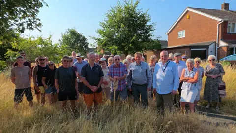 A group of people looking upset stood in long, dry grass, in front of houses. 