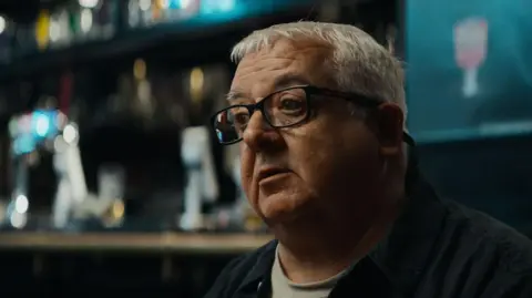 Eight Engines A close-up short of a man with silver hair and black glasses sitting in a bar.