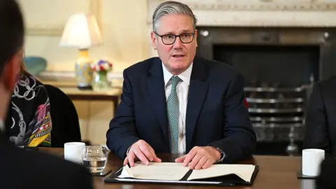 PA Media Sir Keir Starmer sitting at a table speaking at a meeting speaking in 10 Downing Street. He is wearing a green tie has his hands on a note pad on the table 