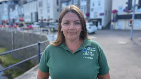 A women with brown hair looking at the camera. She is wearing a green polo short with has the Guernsey Blind Association logo, 'O&M INSTRUCTOR' and 'CHANTELLE' on the left hand side.